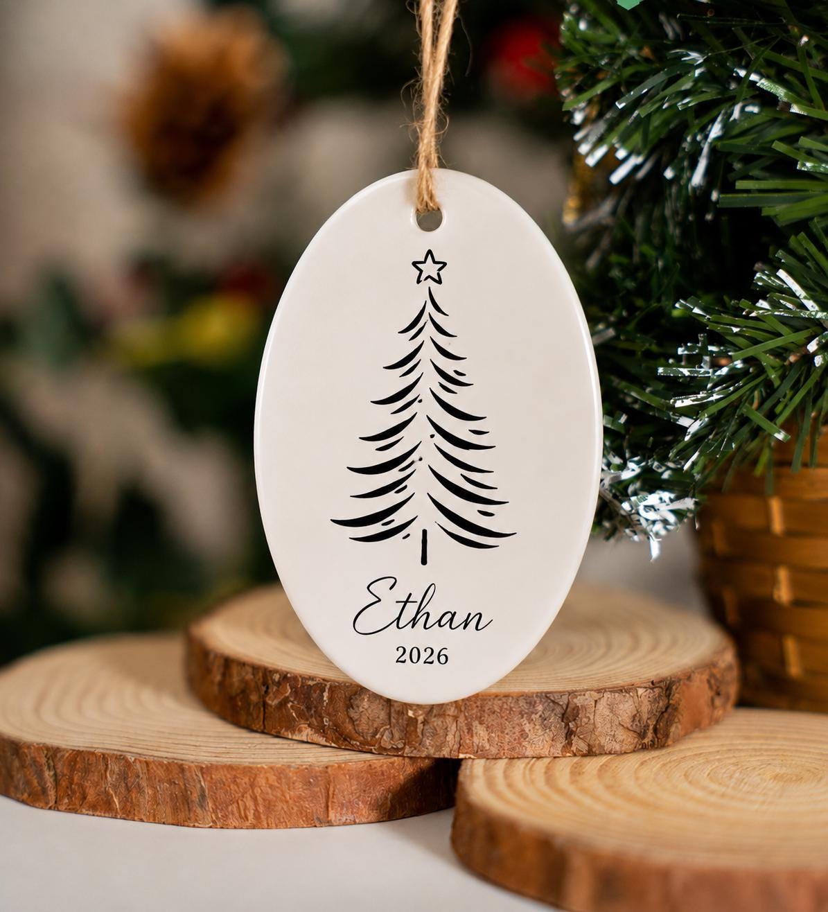Oval ceramic ornament with a sketched pine tree and Ethan 2026 on a twine loop beside wood slices