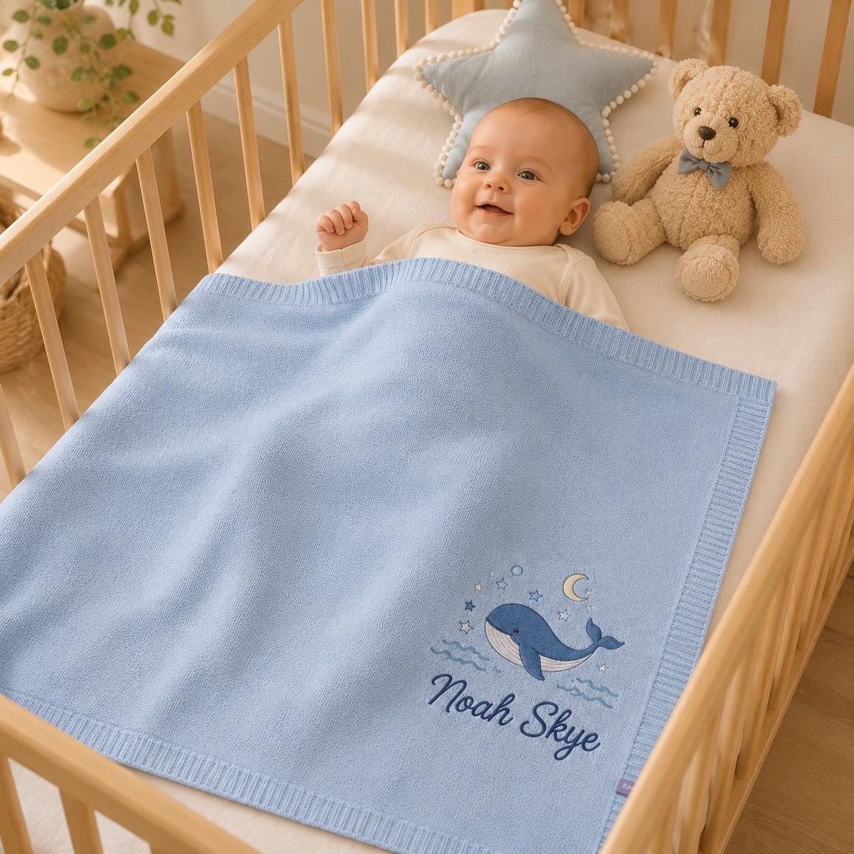 Smiling baby under a blue personalized whale blanket in a crib with a star pillow and a teddy bear