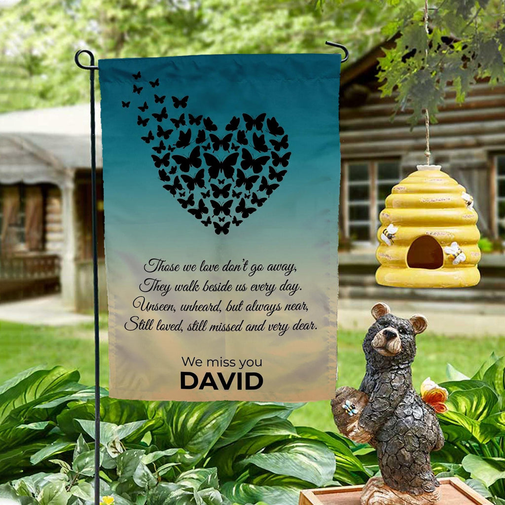 Butterfly heart Those We Love Don't Go Away memorial garden flag on a garden stand by a log cabin with a beehive and bear statue