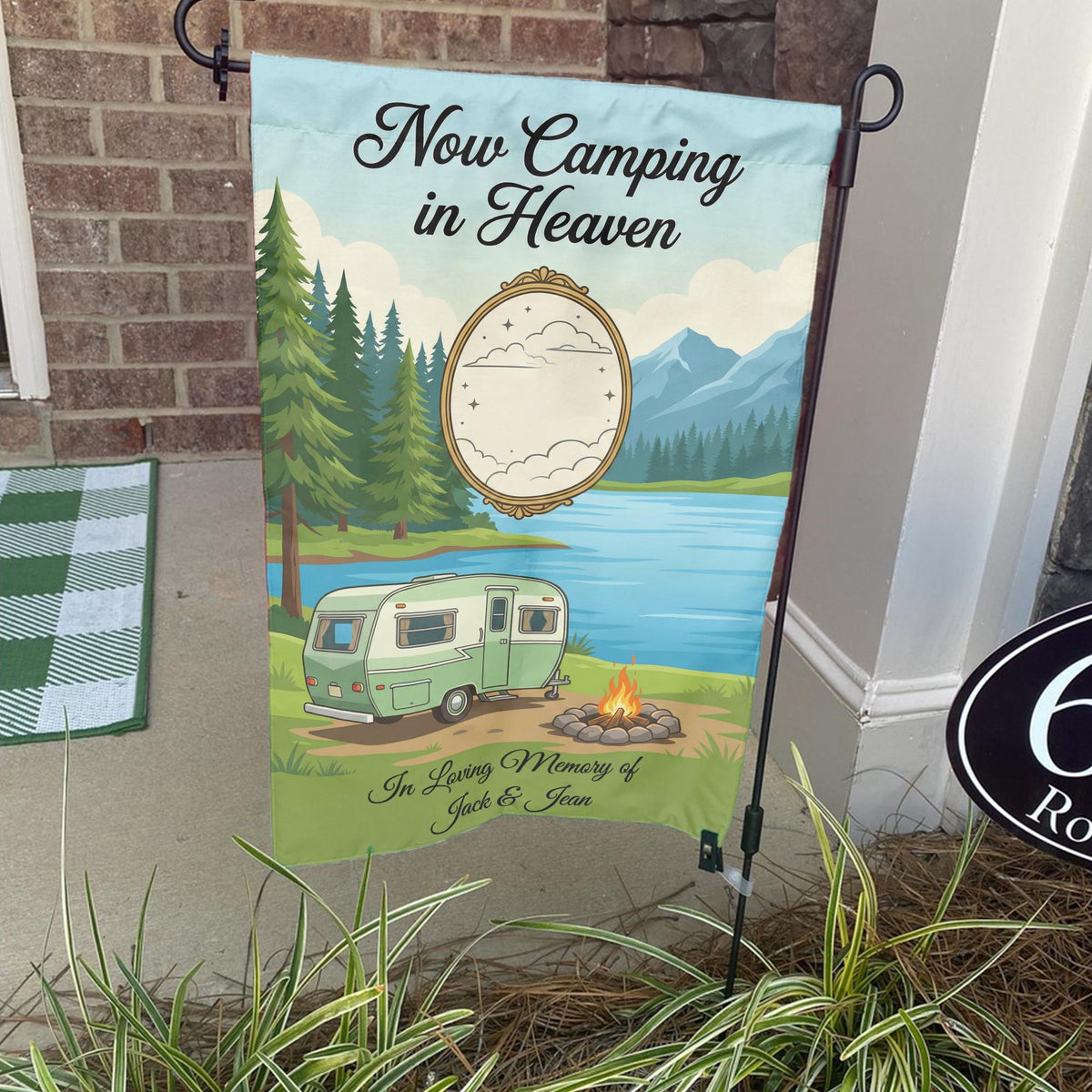 Now Camping in Heaven memorial garden flag with a vintage camper by a mountain lake on a stand by a brick front porch