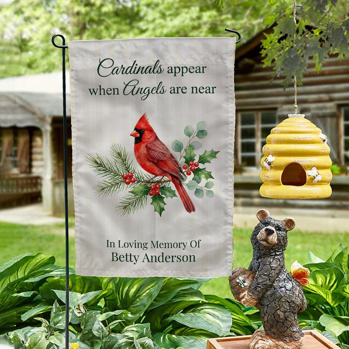 Cardinals Appear When Angels Are Near memorial garden flag with name on a garden stand by a log cabin with a beehive and bear statue
