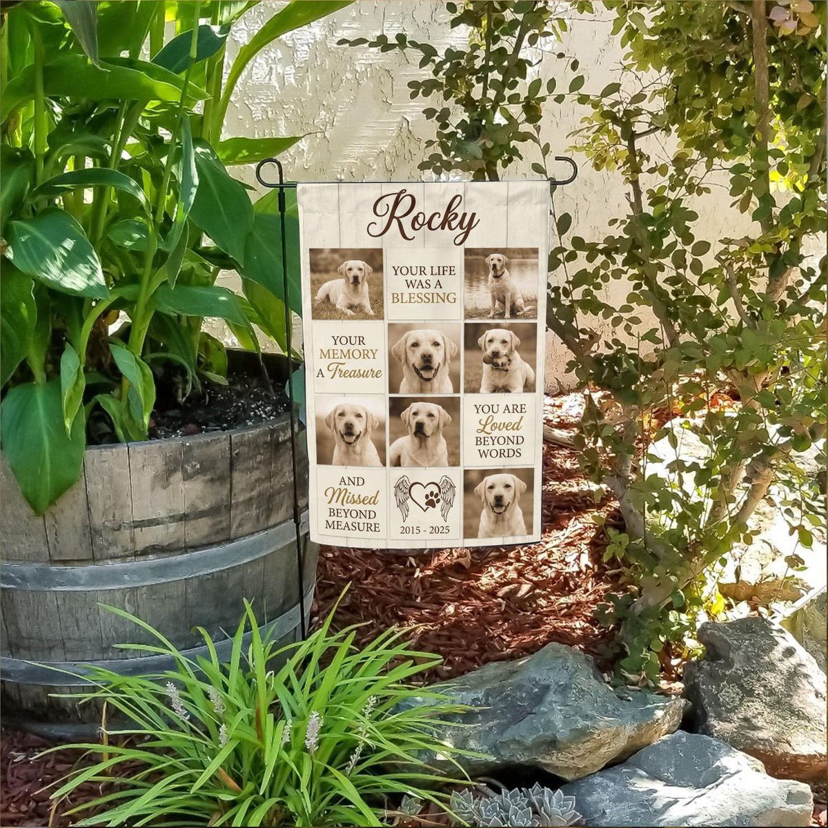 Rustic dog memorial photo collage garden flag with name and years in a flower bed beside a wooden barrel planter