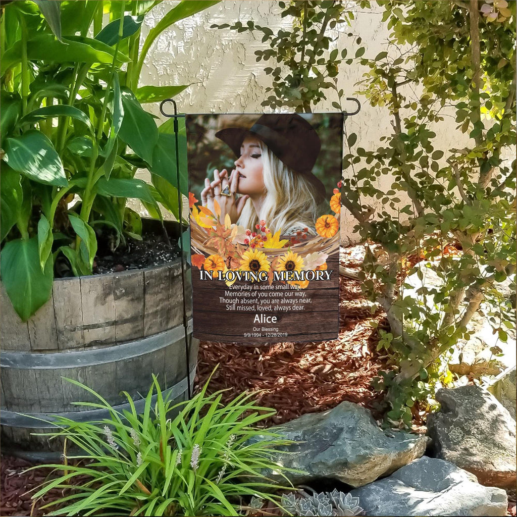 Fall memorial photo garden flag with sunflowers, poem and name in a flower bed beside a wooden barrel planter