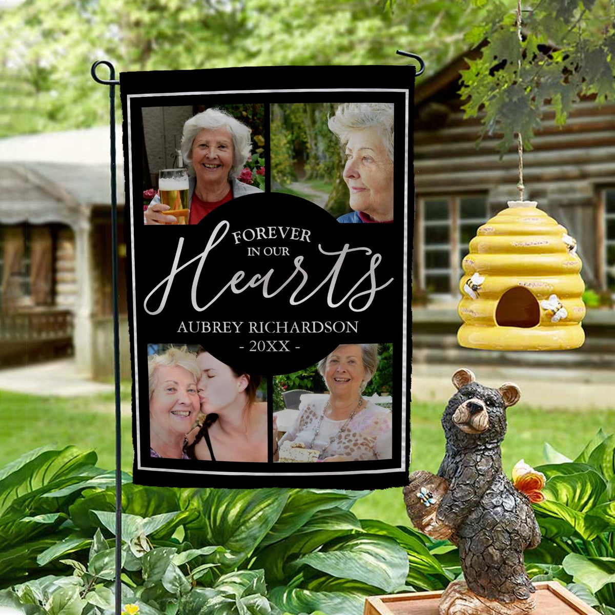 Forever In Our Hearts four photo memorial garden flag with name and year on a garden stand by a log cabin with a beehive and bear statue