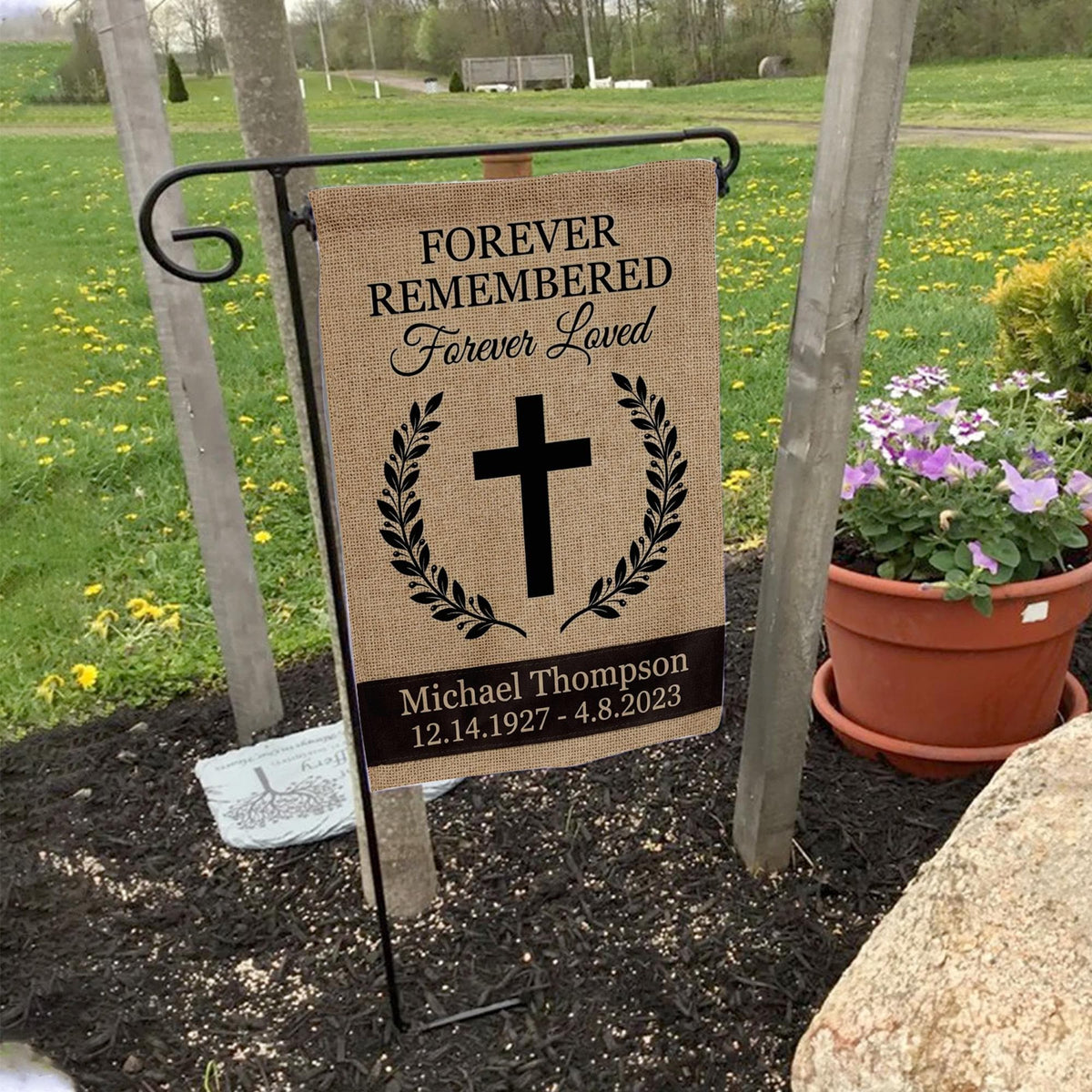 Forever Remembered Forever Loved cross memorial garden flag with burlap look on a garden flag stand in a yard with potted flowers