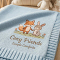 Blue knit baby blanket with an embroidered fox and bunny holding hands above Cozy Friends script lettering