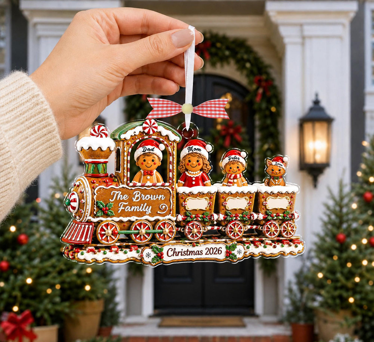 Hand holding a gingerbread train family ornament in front of a decorated front door