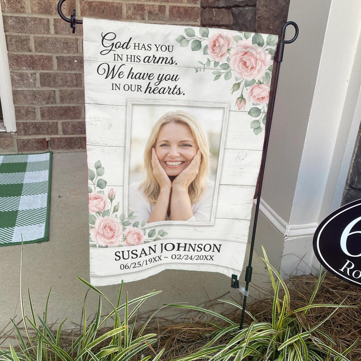 God Has You In His Arms pink rose photo memorial garden flag on a stand by a brick front porch