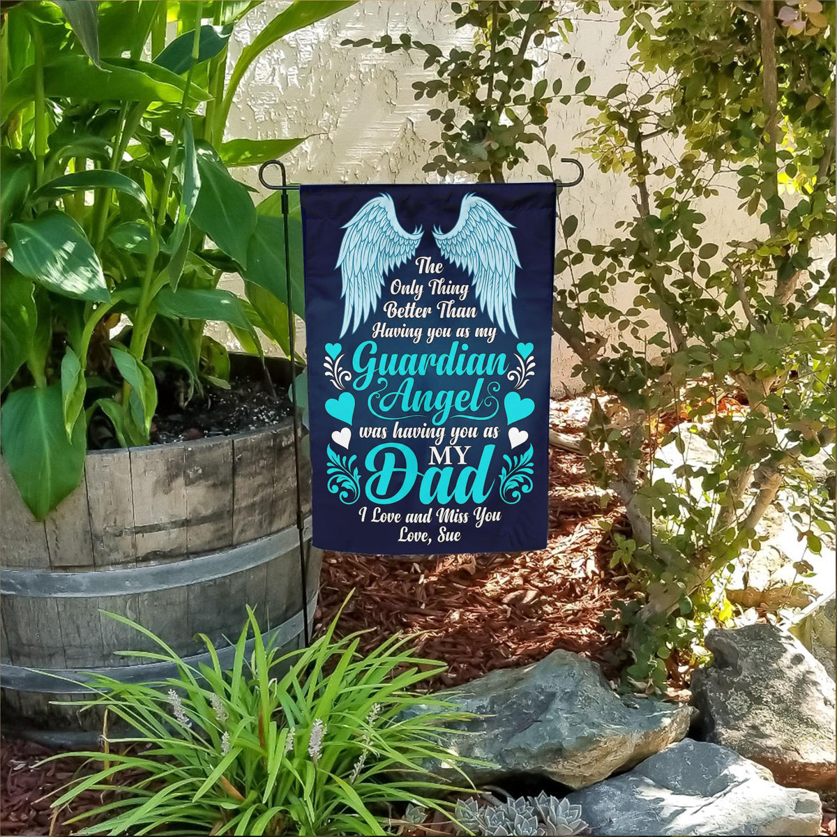 Guardian Angel Dad teal angel wings memorial garden flag in a flower bed beside a wooden barrel planter