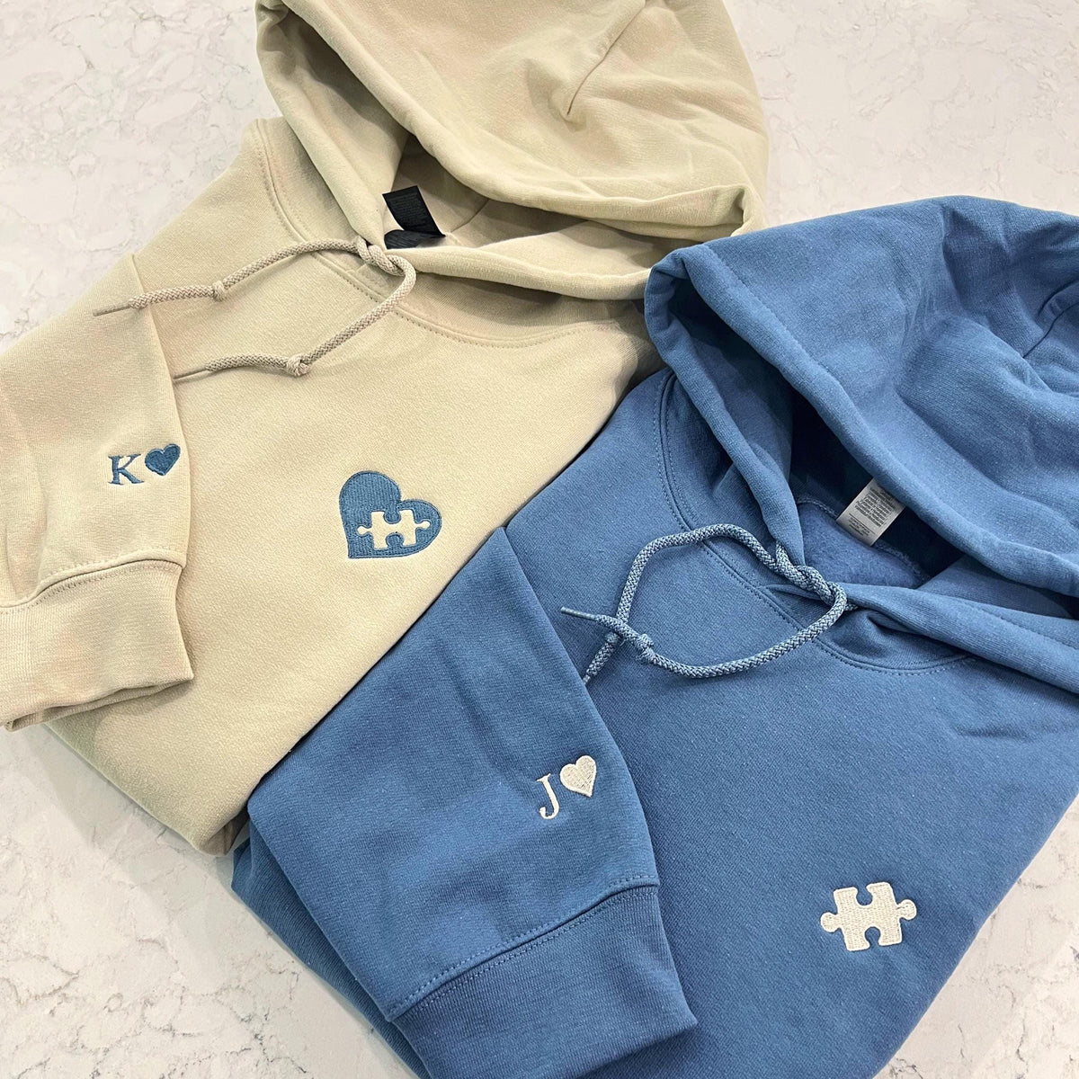 Flat lay of sand and blue couple hoodies with heart and puzzle piece embroidery