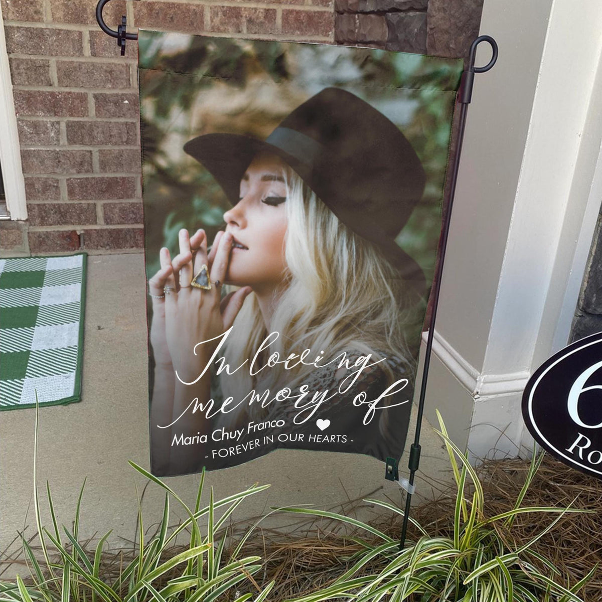 In loving memory photo garden flag with a portrait and name on a stand by a brick front porch