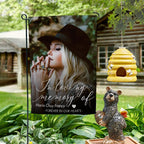 In loving memory photo garden flag with a portrait and name on a garden stand by a log cabin with a beehive and bear statue