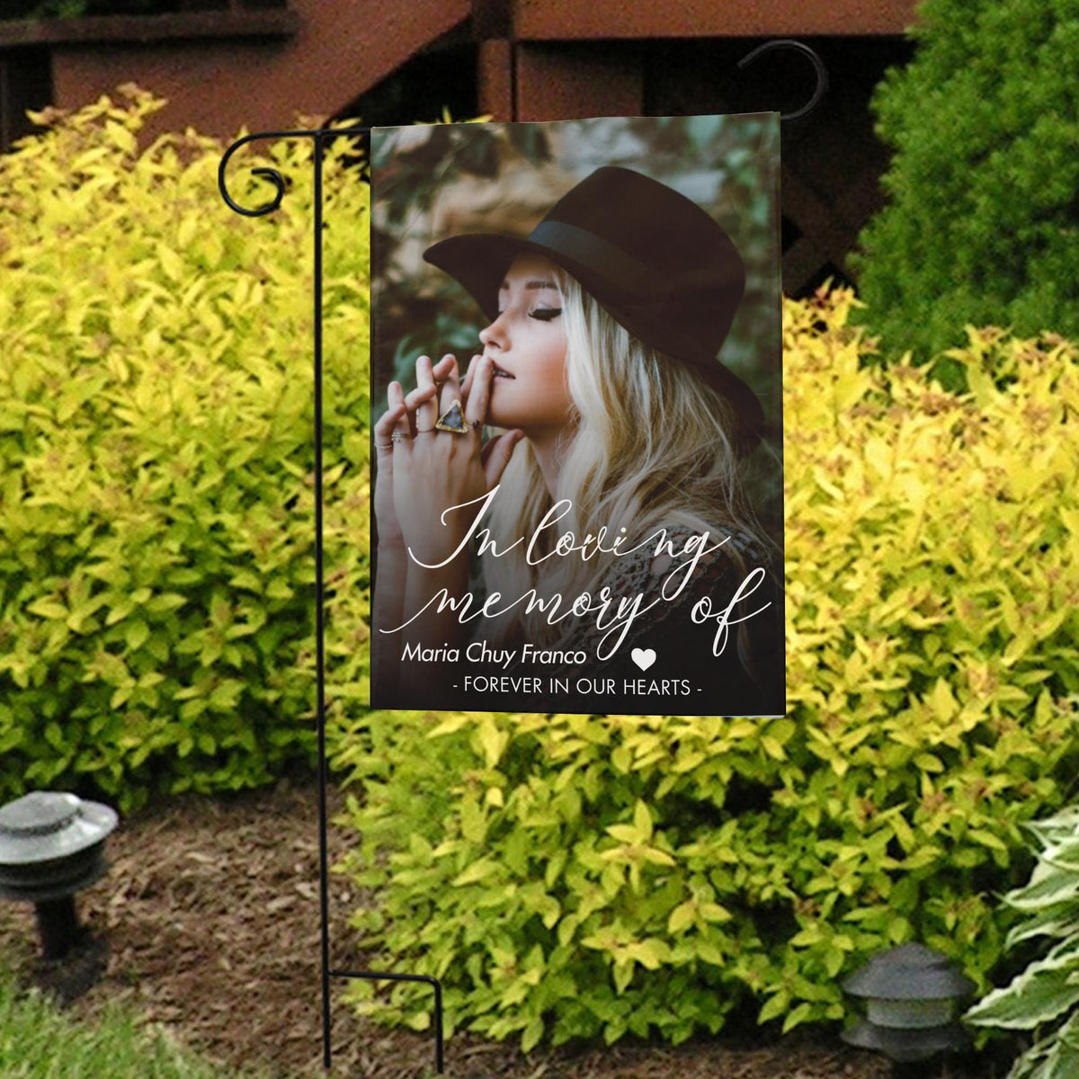 In loving memory photo garden flag with a portrait and name in front of bright yellow shrubs