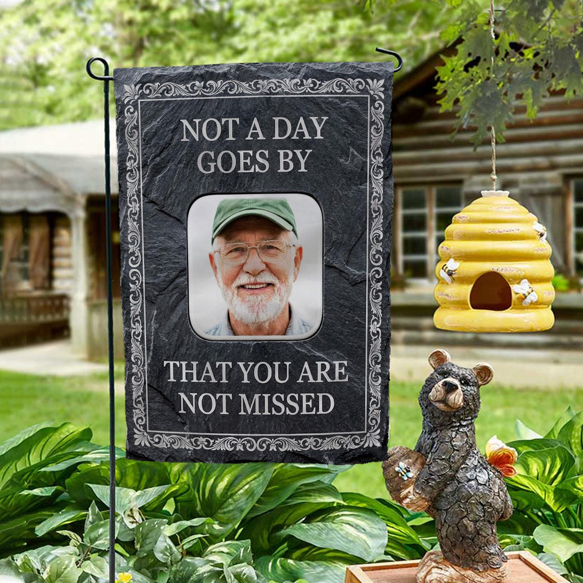 Not A Day Goes By slate style photo memorial garden flag on a garden stand by a log cabin with a beehive and bear statue