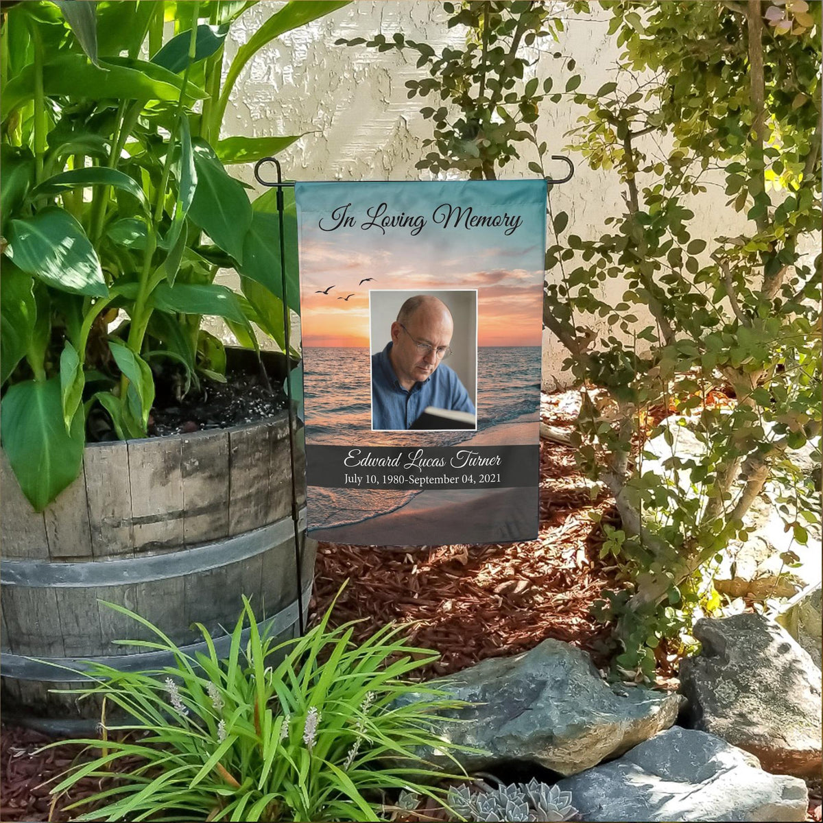 In Loving Memory ocean sunset photo memorial garden flag with name and dates in a flower bed beside a wooden barrel planter