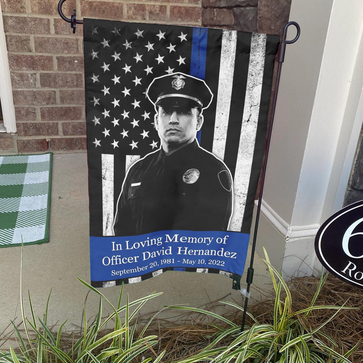 Thin blue line police memorial photo garden flag with officer name and dates on a stand by a brick front porch