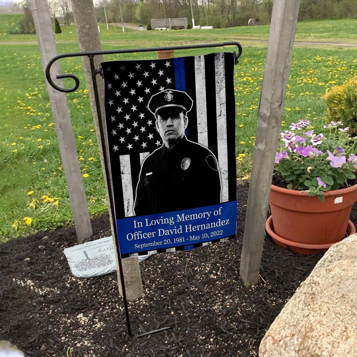 Thin blue line police memorial photo garden flag with officer name and dates on a garden flag stand in a yard with potted flowers