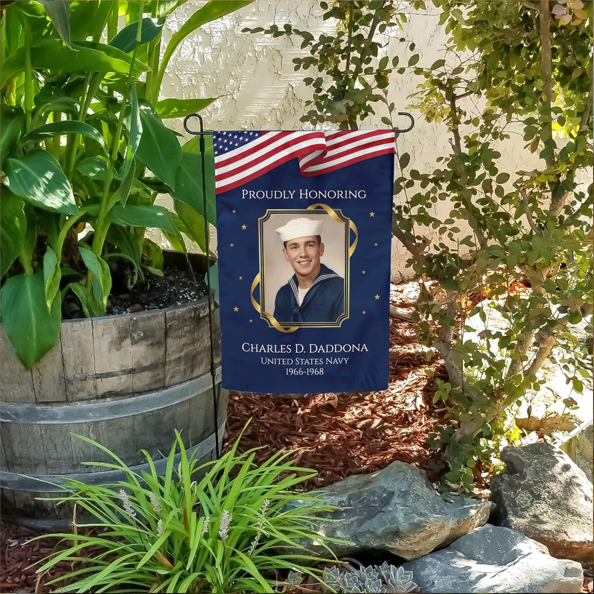 Proudly Honoring military veteran photo memorial garden flag with American flag banner in a flower bed beside a wooden barrel planter