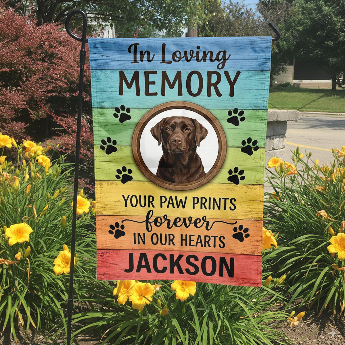Rainbow Your Paw Prints Forever In Our Hearts dog memorial garden flag in a garden bed with yellow lilies