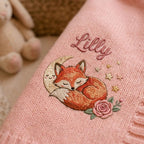 Close-up of an embroidered sleeping fox on a crescent moon with gold stars, a rose and a script name on pink knit