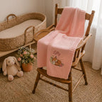Pink knit baby blanket with fox and moon embroidery and a script name draped over a wooden chair in a nursery