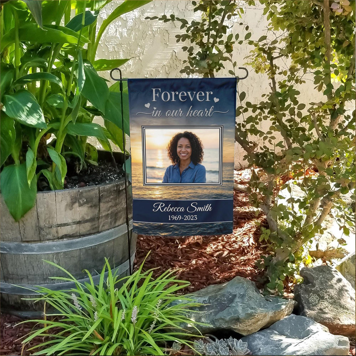 Forever In Our Heart sunset beach photo memorial garden flag with name and years in a flower bed beside a wooden barrel planter