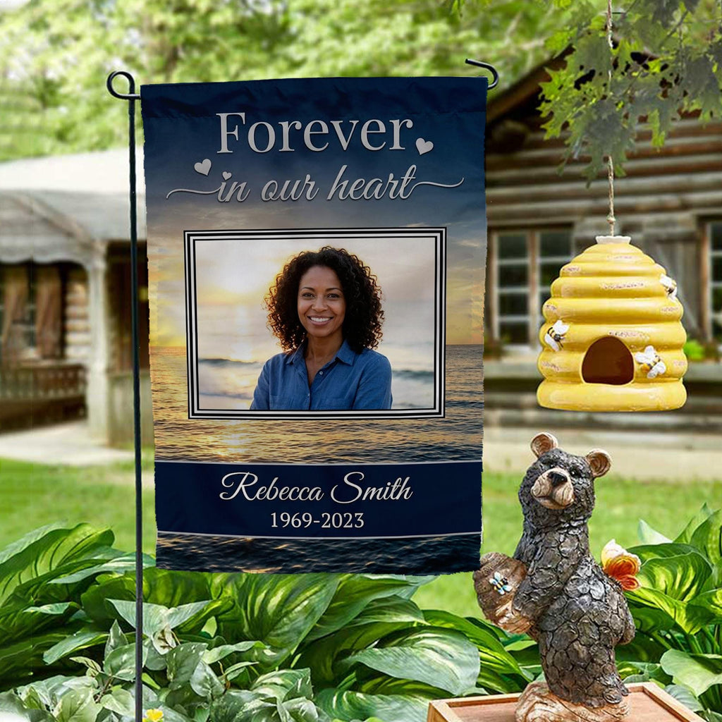 Forever In Our Heart sunset beach photo memorial garden flag with name and years on a garden stand by a log cabin with a beehive and bear statue
