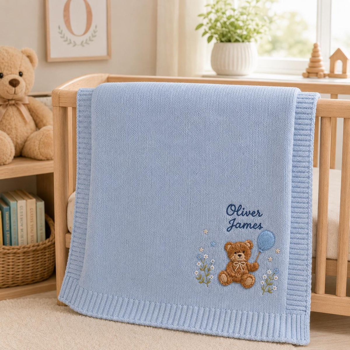 Blue personalized teddy bear balloon baby blanket draped over a wood crib beside shelves with books and a bear