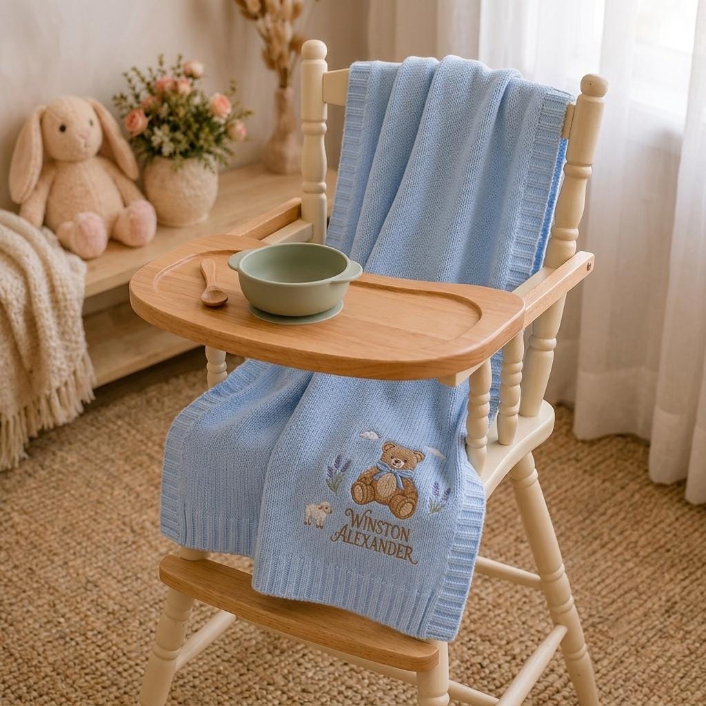 Blue teddy bear name blanket with lamb and lavender embroidery draped over a wooden high chair in a nursery