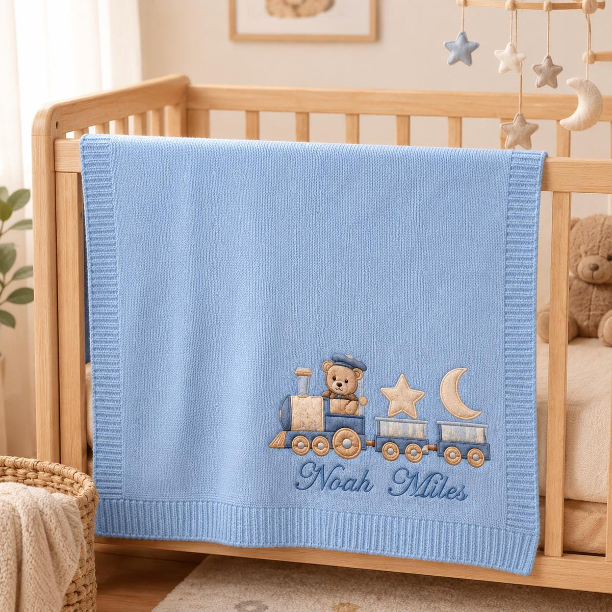Personalized blue train baby blanket hanging over the side of a natural wood crib in a cozy nursery
