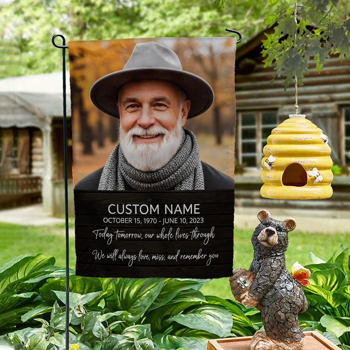 Today Tomorrow Always portrait memorial garden flag with name and dates on a garden stand by a log cabin with a beehive and bear statue