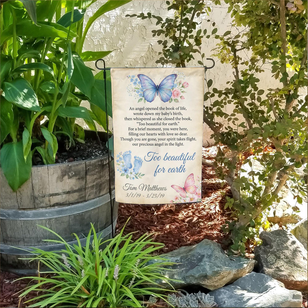 Too Beautiful For Earth butterfly baby memorial garden flag with name and dates in a flower bed beside a wooden barrel planter
