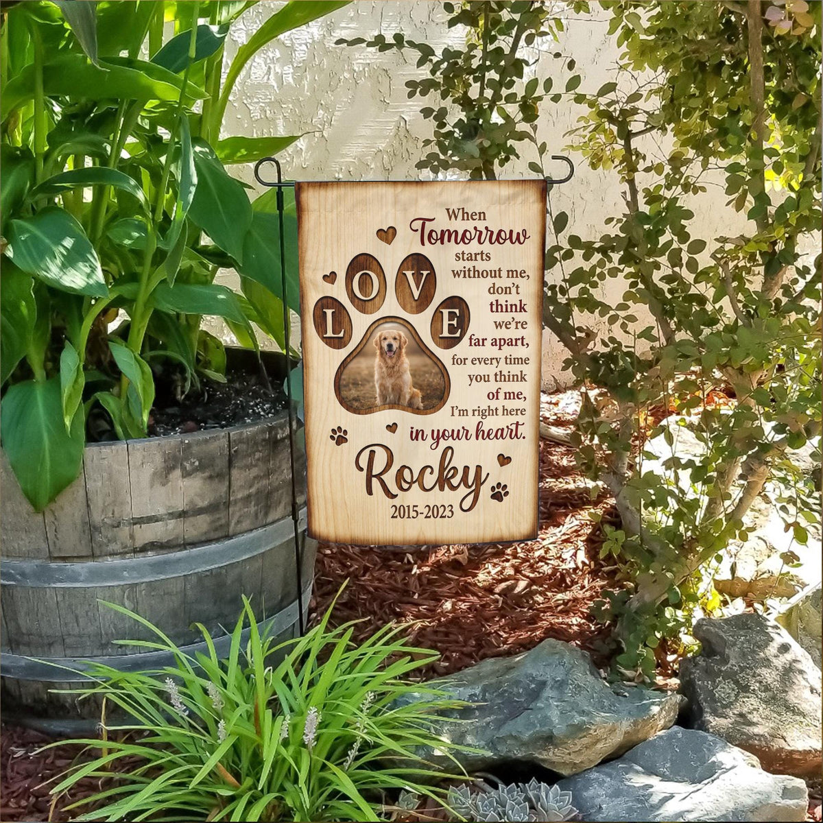 When Tomorrow Starts Without Me dog memorial garden flag with paw print photo in a flower bed beside a wooden barrel planter
