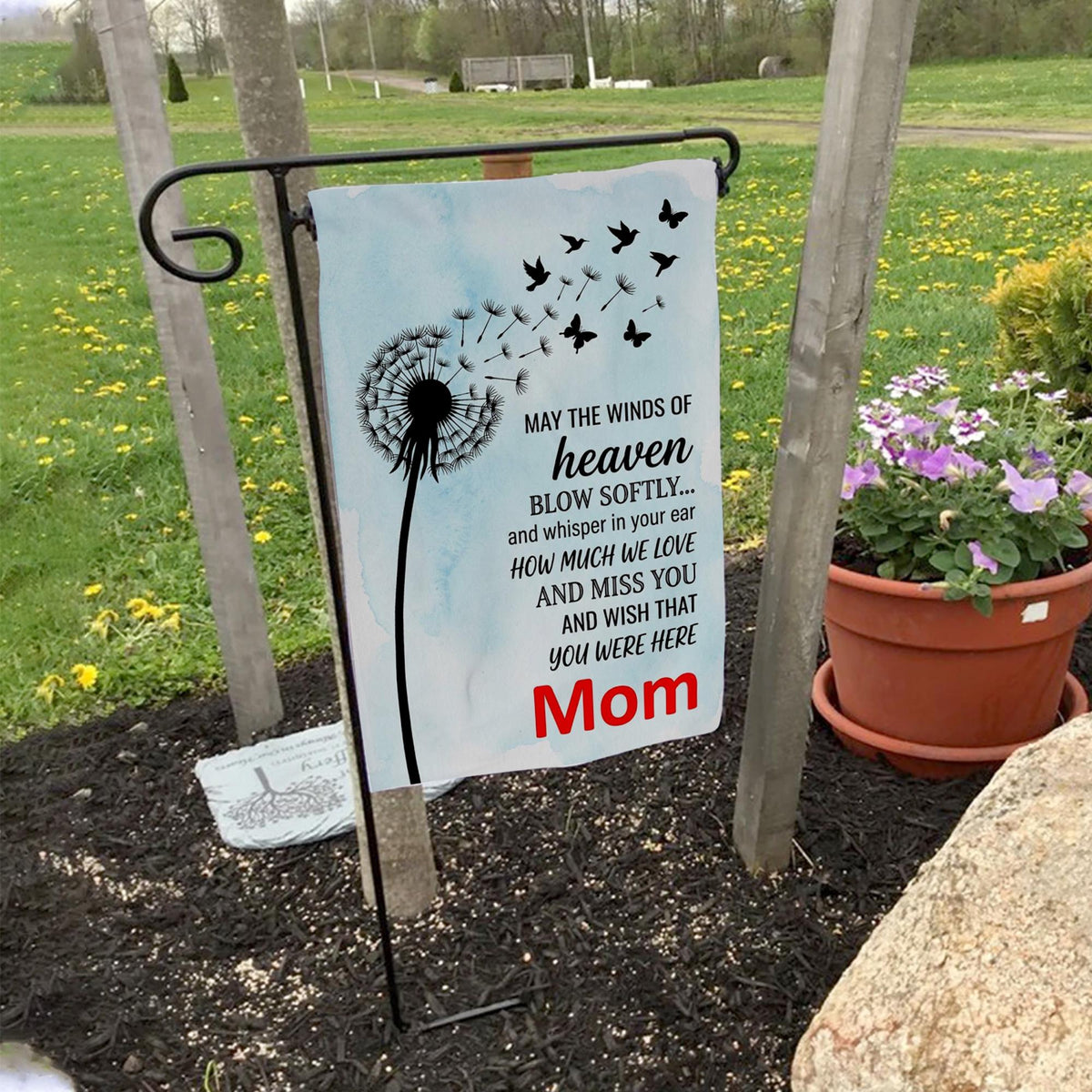 May the Winds of Heaven dandelion memorial garden flag for Mom on a garden flag stand in a yard with potted flowers